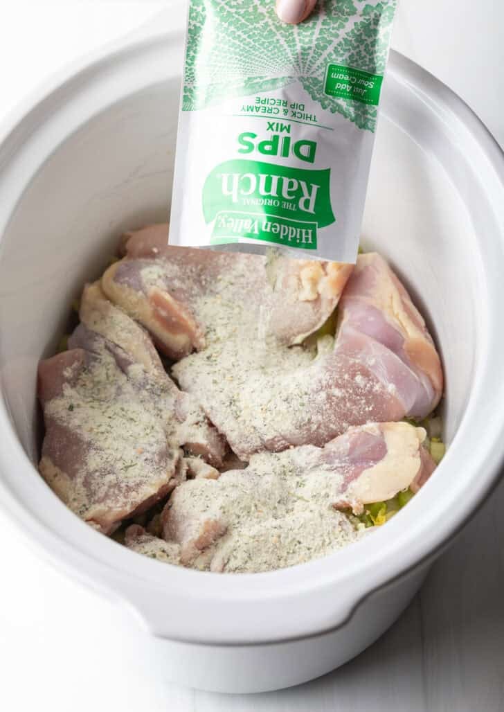 Sprinkling ranch dip powder from the packet over a white crockpot full of boneless skinless chicken breasts.