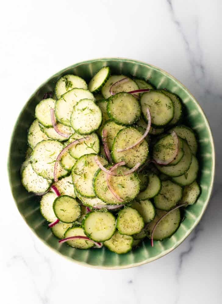Top down view green plate loaded with sliced cucumbers and red onion salad with dill.