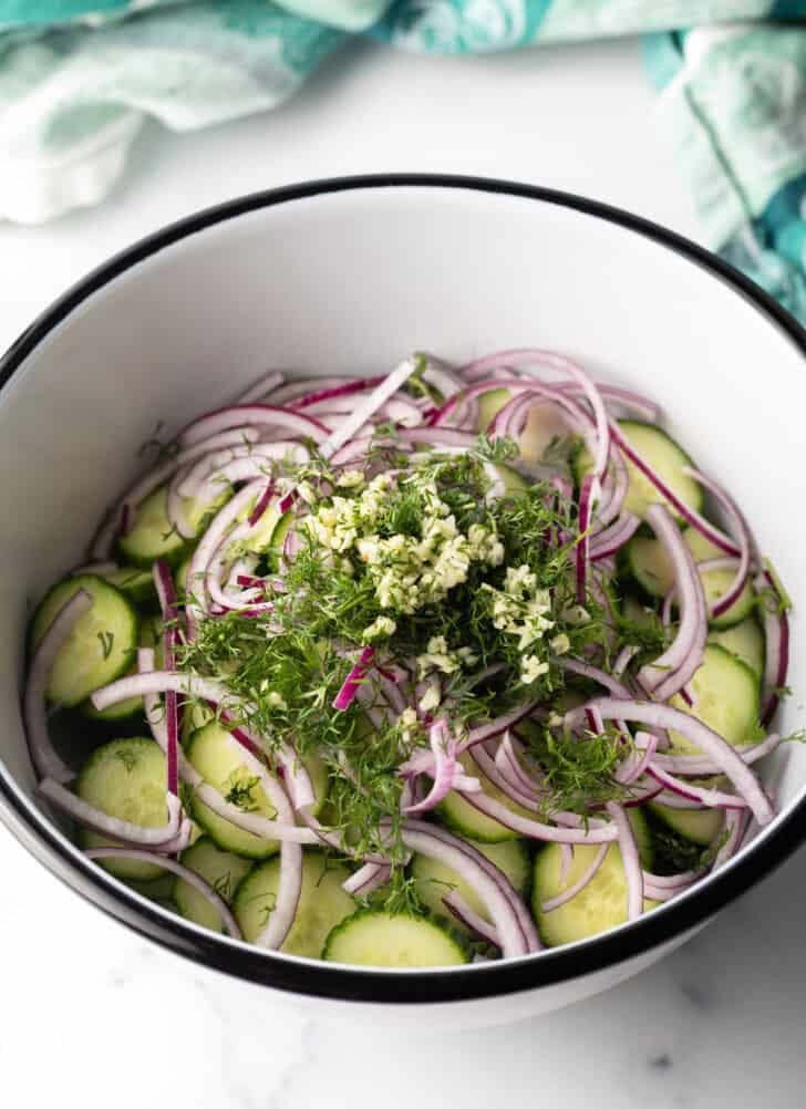 White bowl with slices of cucumbers, red onions, chopped dill, and minced garlic.