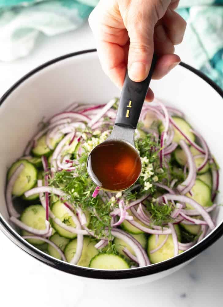 Hand adding vinegar from a tablespoon into a bowl with sliced cucumbers, red onions, and dill