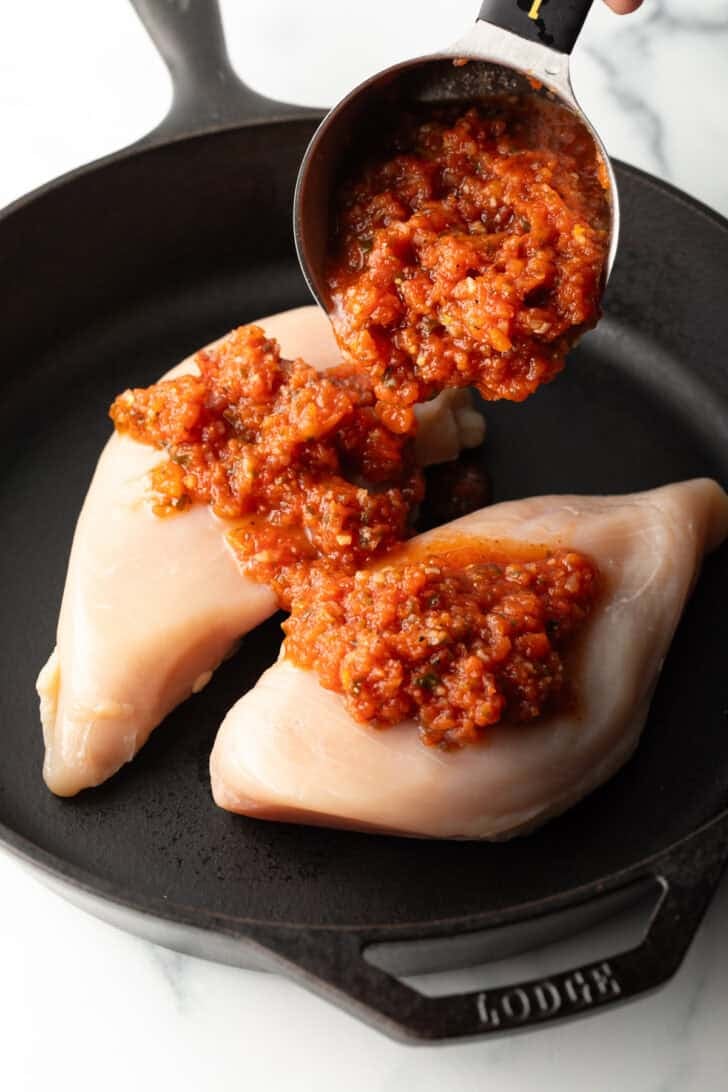 Adding tomato salsa from a measuring cup onto two raw chicken breasts in a cast iron skillet.