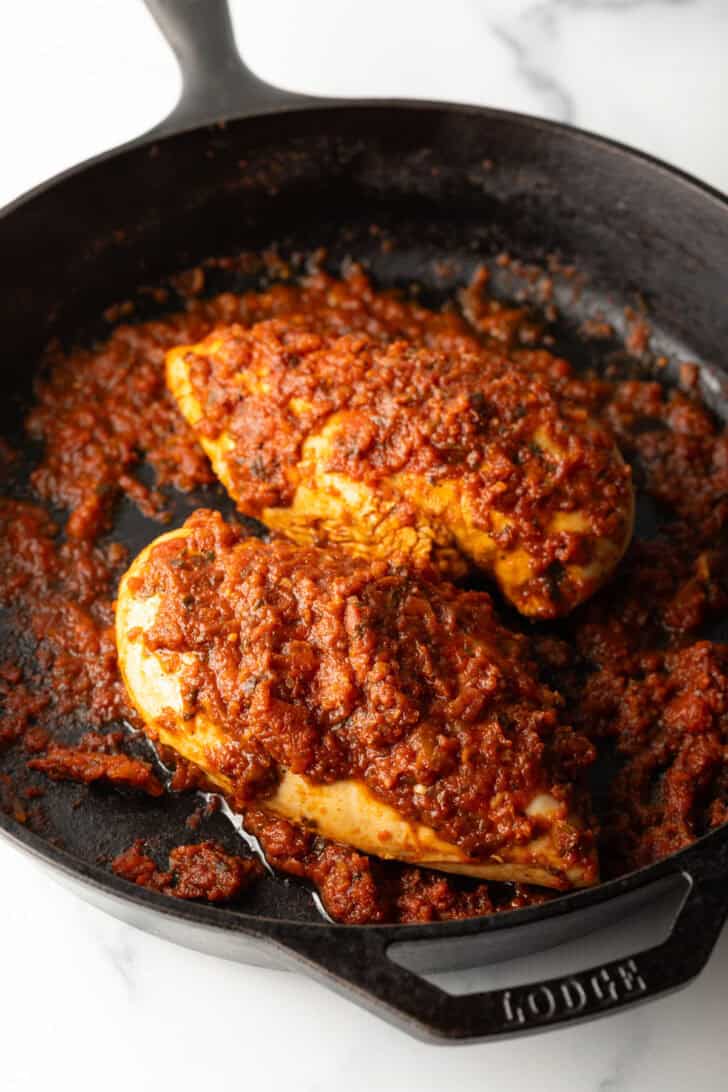 Two chicken breasts in a cast iron skillet with tomato sauce.