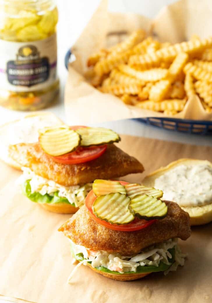 Two open face fried fish sandwiches, each topped with a slice of tomato and pickle slices.