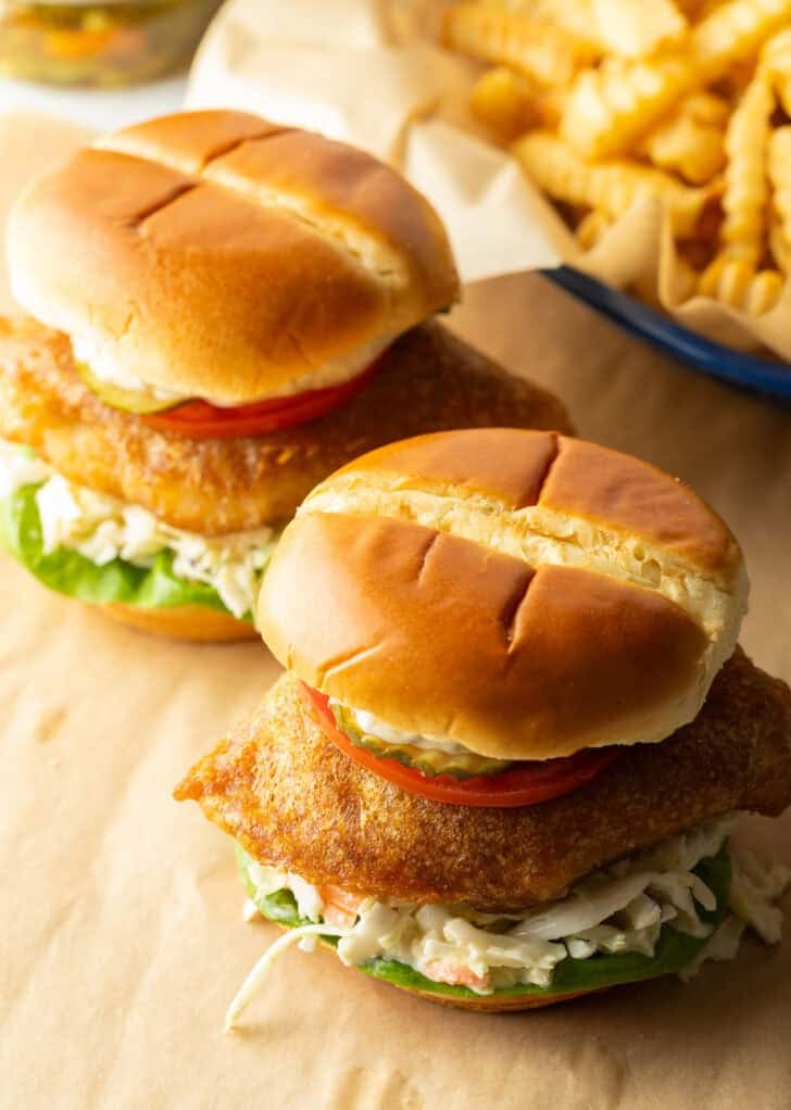 Two battered and fried fish sandwiches on a bun with coleslaw, tartar sauce, and a slice of tomato.