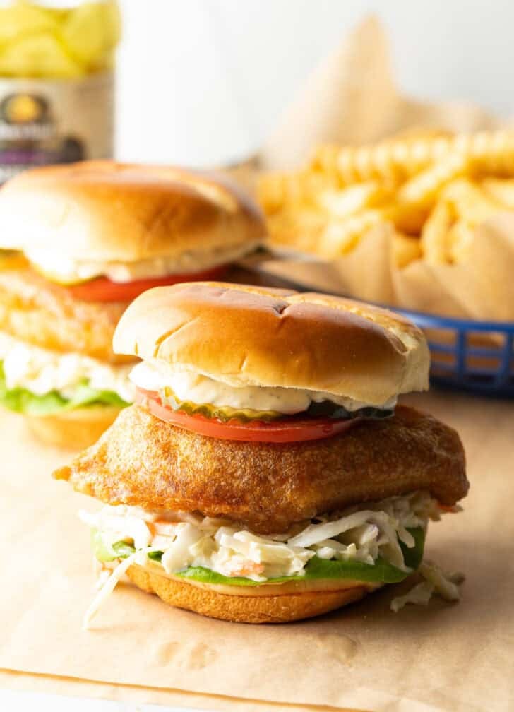 Two battered and fried fish sandwiches on a bun with coleslaw, tartar sauce, and a slice of tomato.