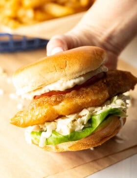 Hand holding a battered and fried fish sandwich on a bun with coleslaw, tartar sauce, and a slice of tomato.