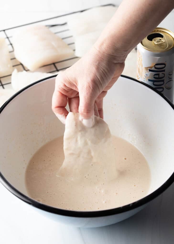Hand dipping a piece of flour-coated fish into batter.