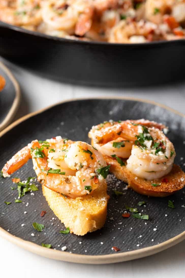 Black oval plate with two baguette slices topped with garlic butter shrimp and herbs.