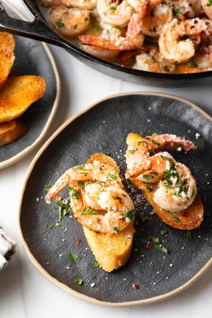 Black oval plate with two baguette slices topped with garlic butter shrimp and herbs.