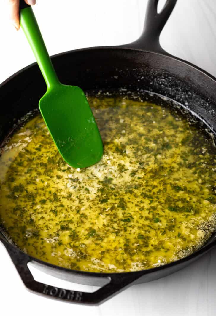 Stirring garlic, butter, and fresh herbs in a large skillet with green spatula.