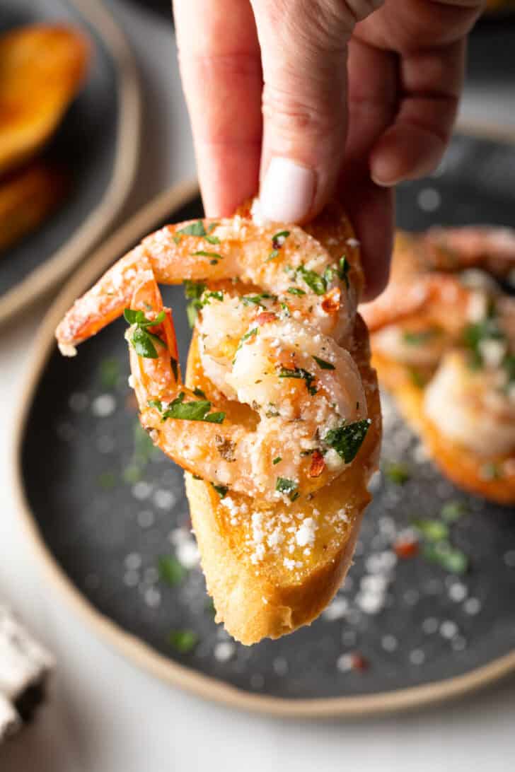 Hand holding a piece of baguette topped with garlic butter shrimp.