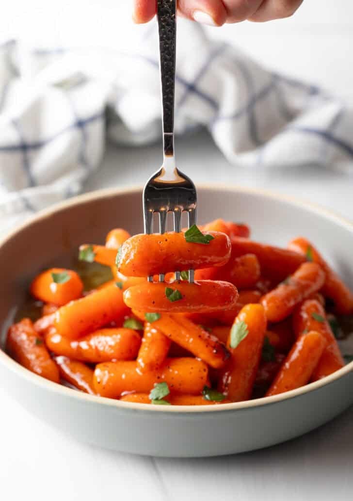 A fork is taking several roasted baby carrots out of a bowl.