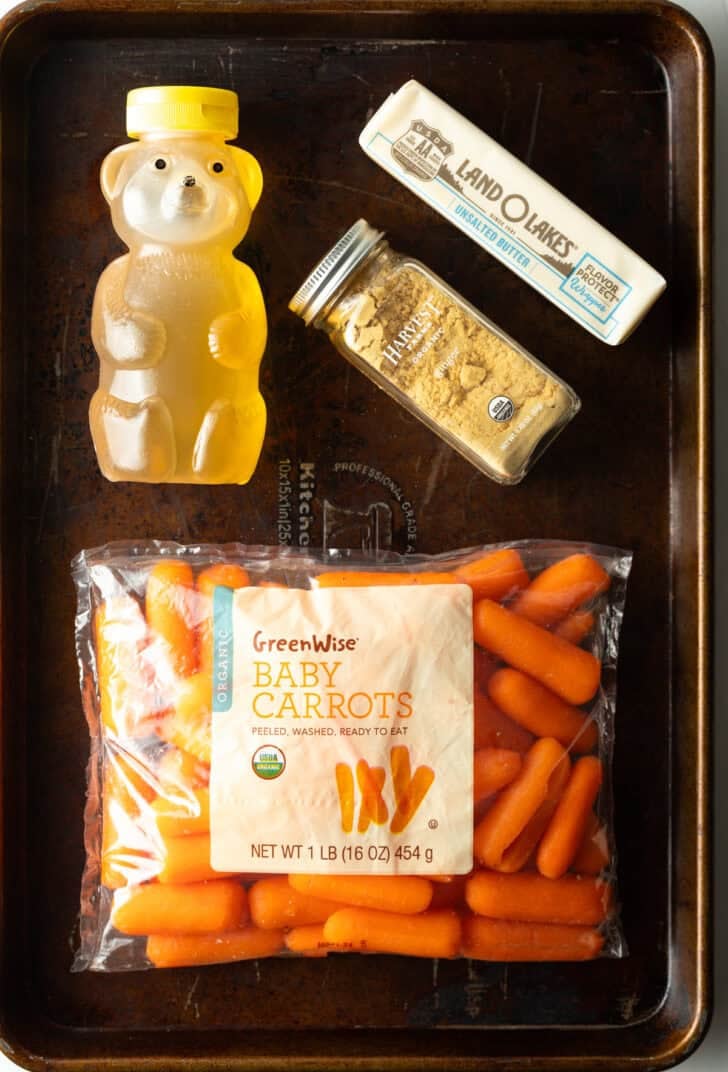 Top down view of ingredients: Bag of baby carrots, bear bottle of honey, garlic powder jar, and stick of butter.