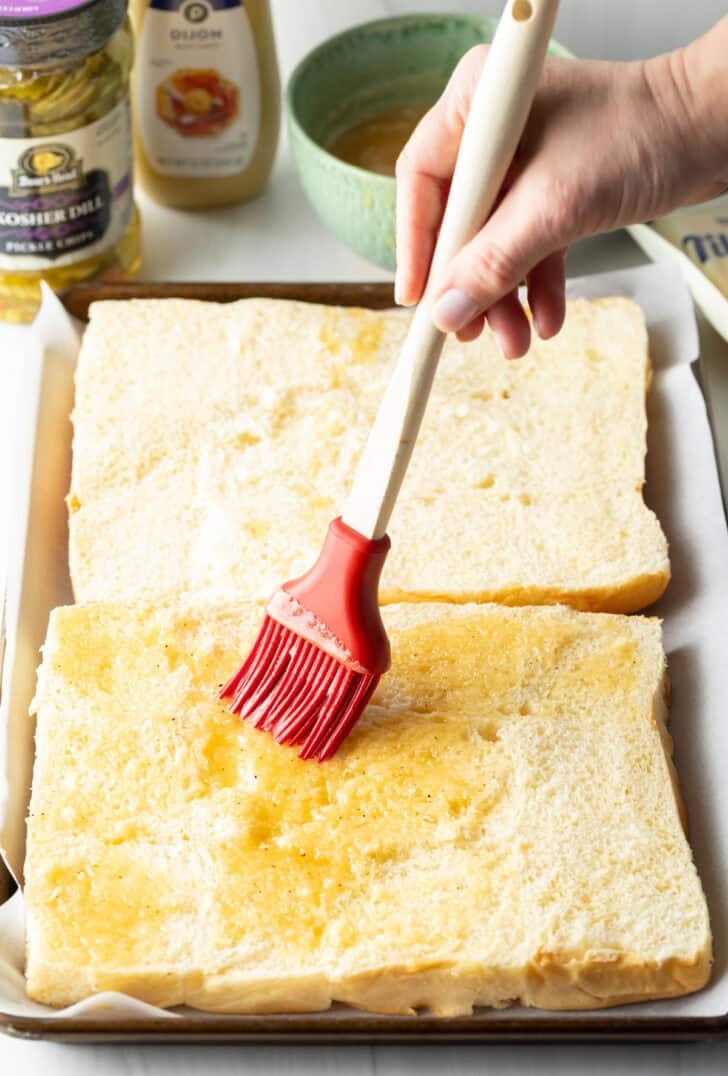 Hand with red silicone brush coating the bottom layer of Hawaiian rolls with butter.