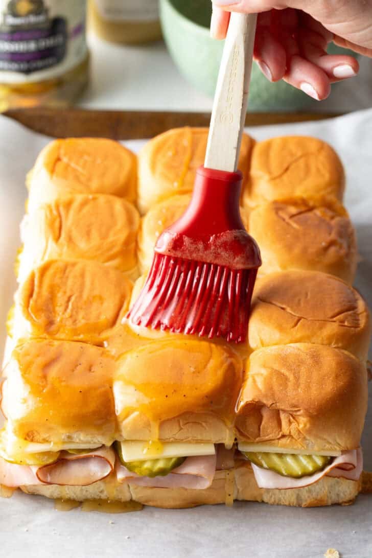 Using a red silicone brush to coat the top of a dozen assembled sliders withe melted butter.