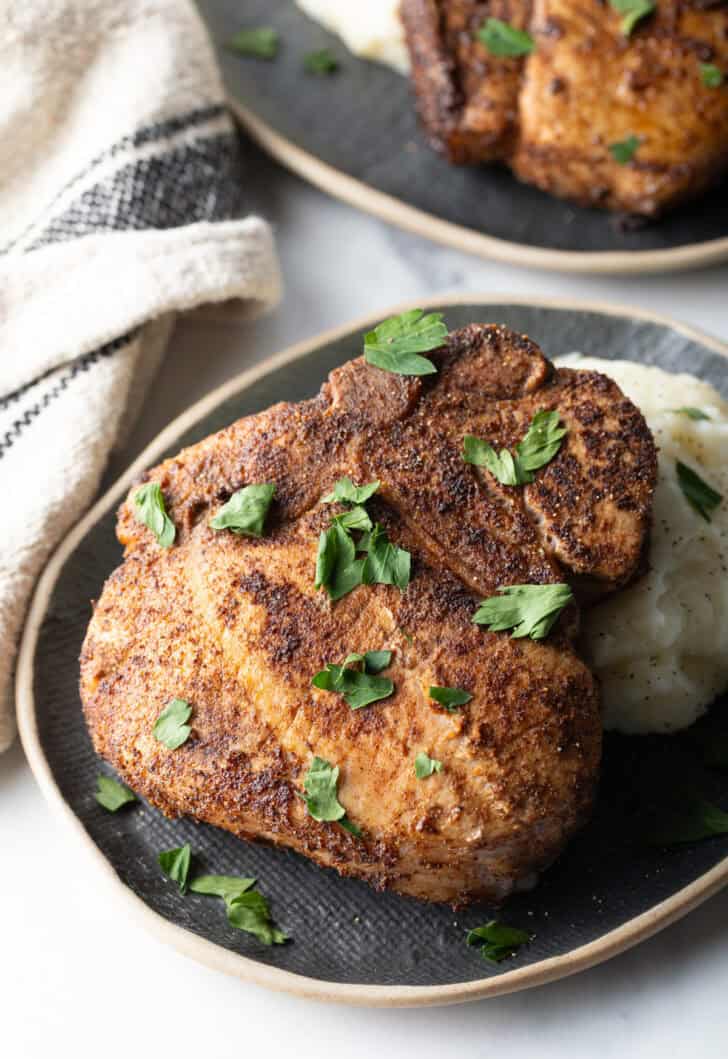 Browned seasoned bone-in pork chop on a plate with mashed potatoes, garnished with chopped parsley.