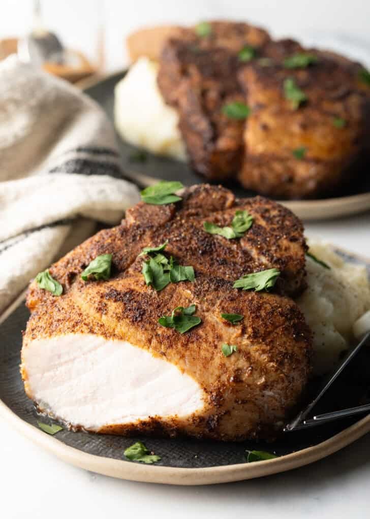 Browned seasoned bone-in pork chop on a plate with mashed potatoes, garnished with chopped parsley. The front piece of the chop has been cut off to show the juice meat inside.