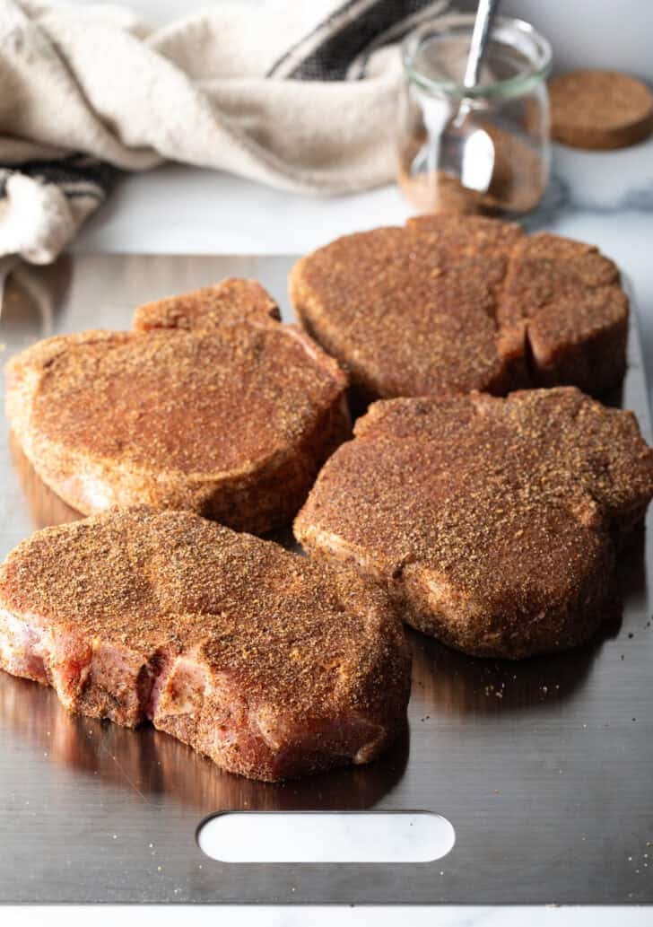 Four raw seasoned bone-in pork chops on a baking sheet.