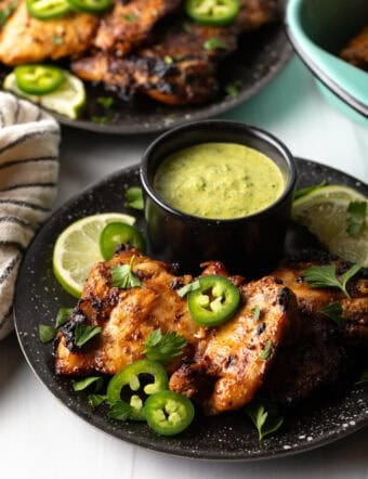 Black plate with Peruvian chicken pieces, sliced jalapenos, chopped cilantro, and lime wedge. There is a black ramekin of green sauce.