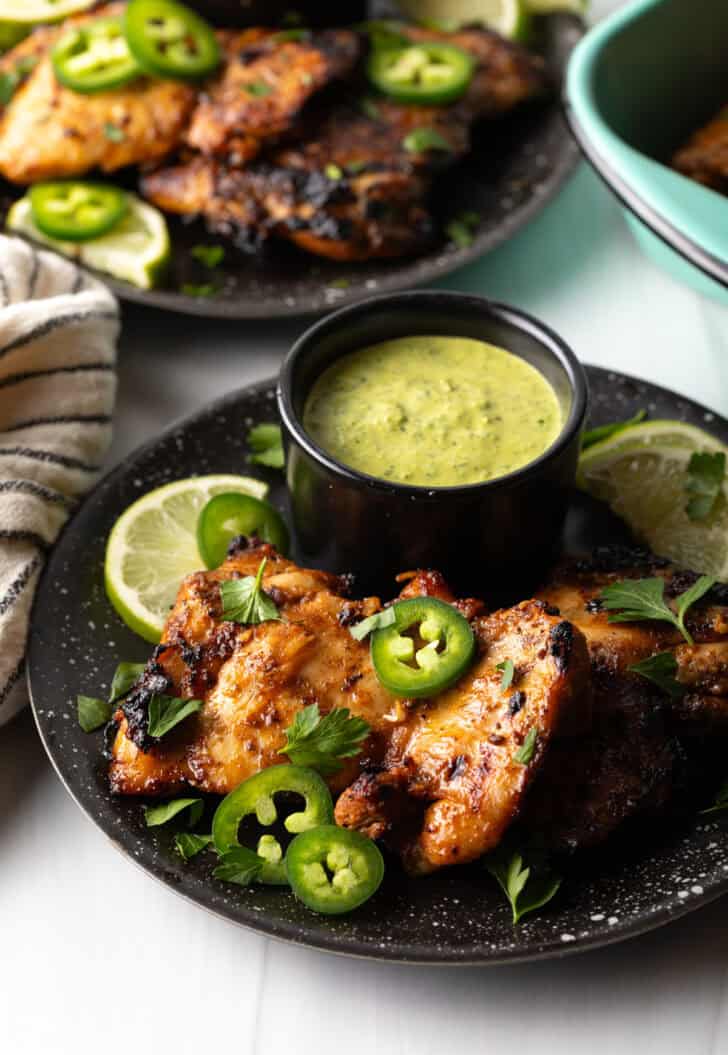Black plate with Peruvian chicken pieces, sliced jalapenos, chopped cilantro, and lime wedge. There is a black ramekin of green sauce.