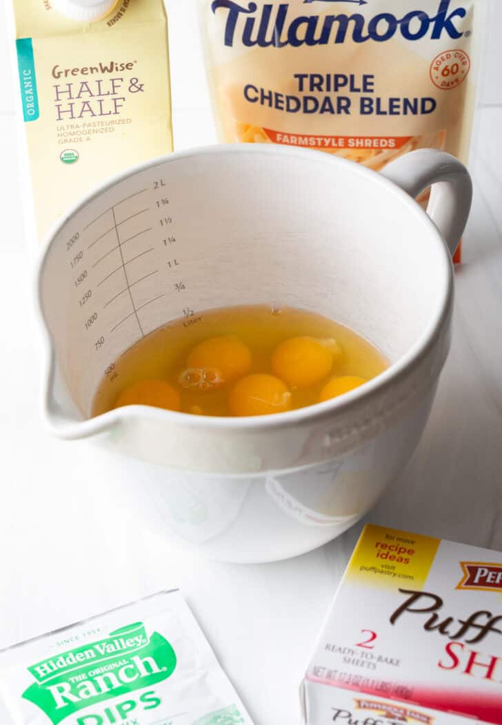 Recipe ingredients: Large white mixing pitcher with several eggs, a box of puff pastry, packet of ranch dressing mix, bag of shredded cheese, and box of half and half.