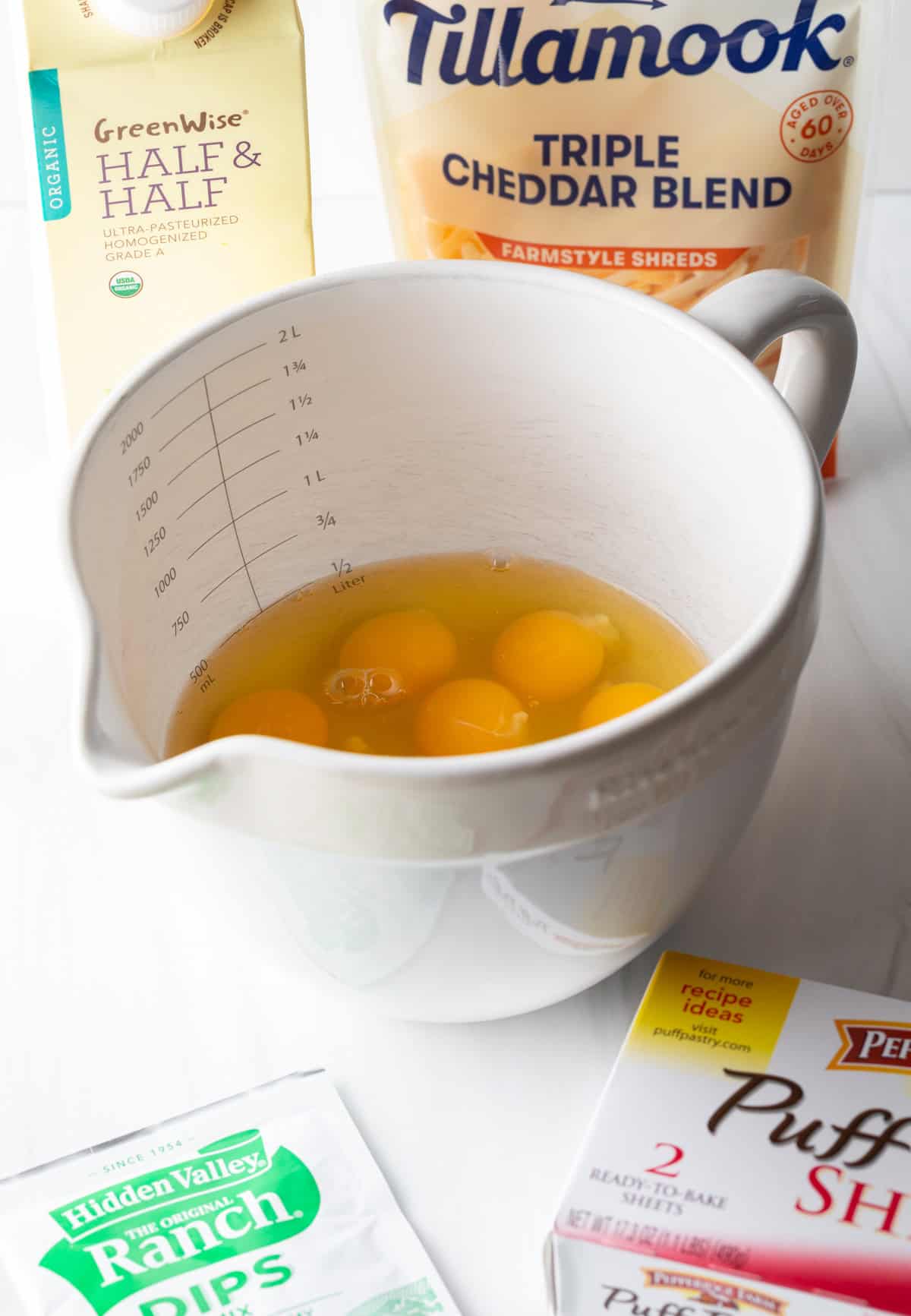 Recipe ingredients: Large white mixing pitcher with several eggs, a box of puff pastry, packet of ranch dressing mix, bag of shredded cheese, and box of half and half.