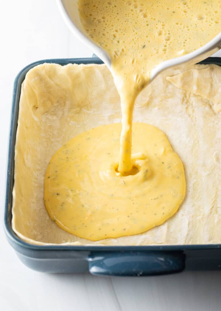 Pouring blended egg mixture from a measuring cup into a prepared baking dish lined with puff pastry.