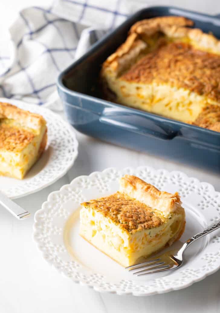 Square of egg casserole on a white plate with a fork. In the background is a square baking dish with the casserole.