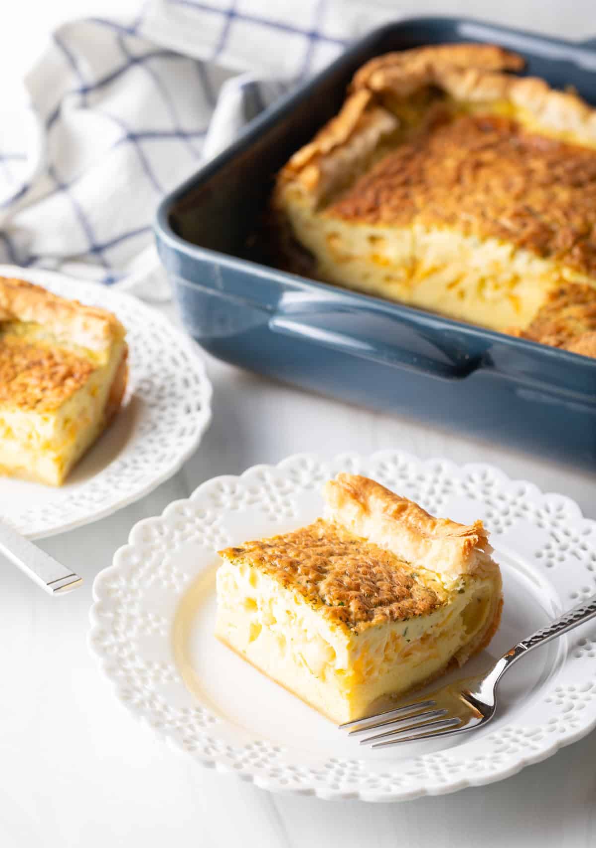 Square of egg casserole on a white plate with a fork. In the background is a square baking dish with the casserole.