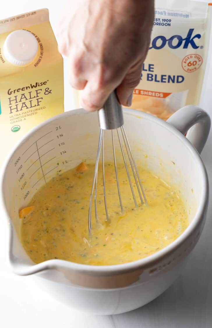 Hand whisking eggs and cheese in a large white mixing pitcher.