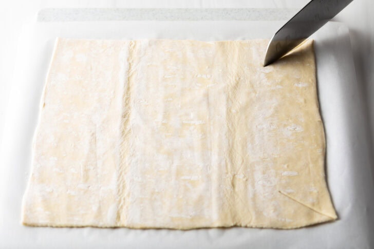 Using a sharp knife to slice a large rectangle of puff pastry.