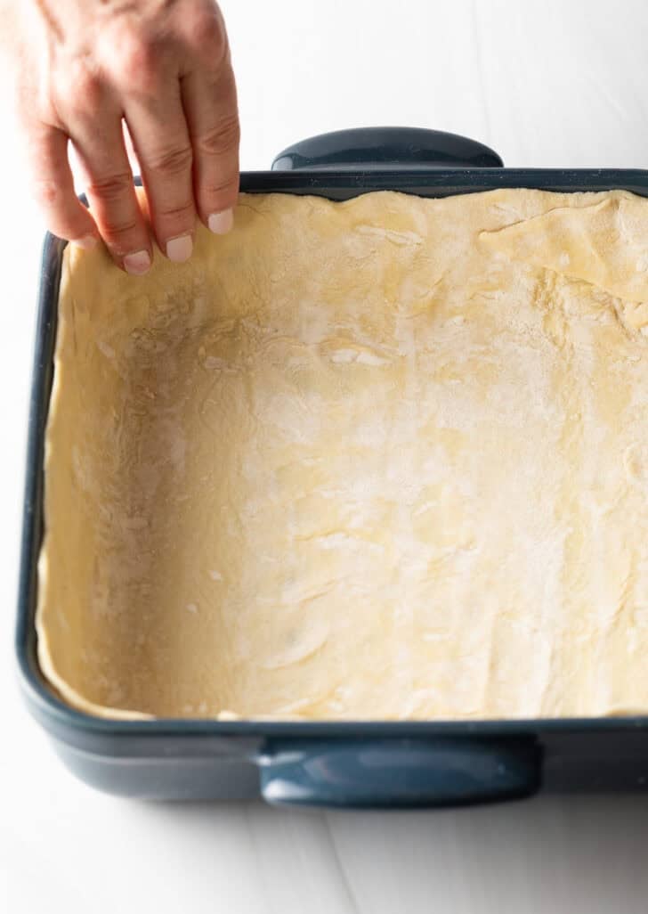 Hand placing pastry dough into the bottom and sides of a square baking dish.
