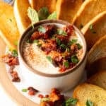 White cup with whipped feta with chopped sun dried tomato, on a plate surrounded by slices of toasted baguette.