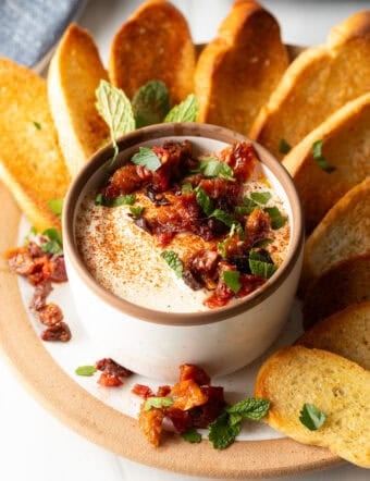 White cup with whipped feta with chopped sun dried tomato, on a plate surrounded by slices of toasted baguette.
