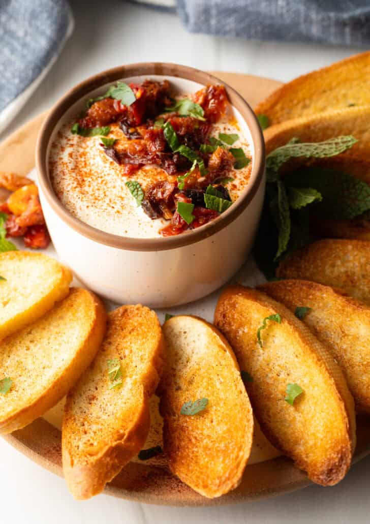 White cup with whipped feta with chopped sun dried tomato, on a plate surrounded by slices of toasted baguette.