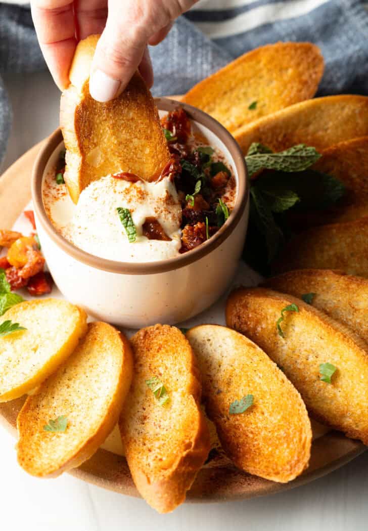 Hand dipping a slice of toasted baguette into a bowl of creamy whipped feta dip.