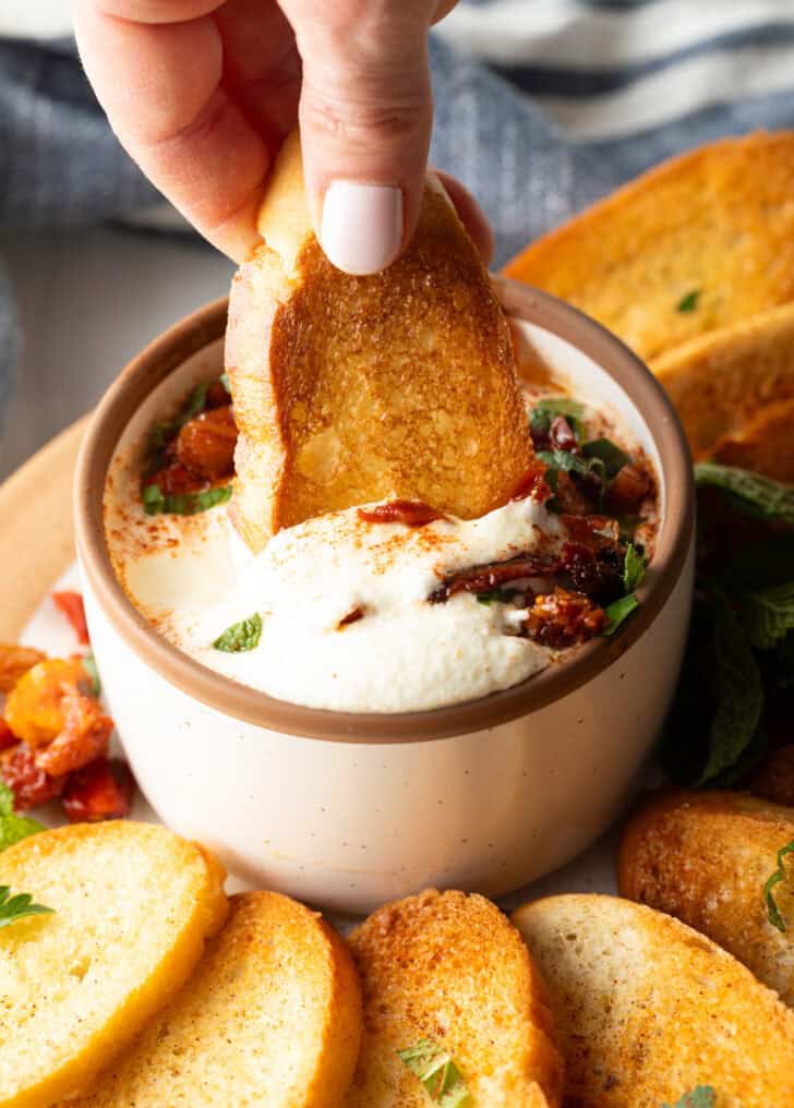 Hand dipping a slice of toasted baguette into a bowl of creamy whipped feta dip.