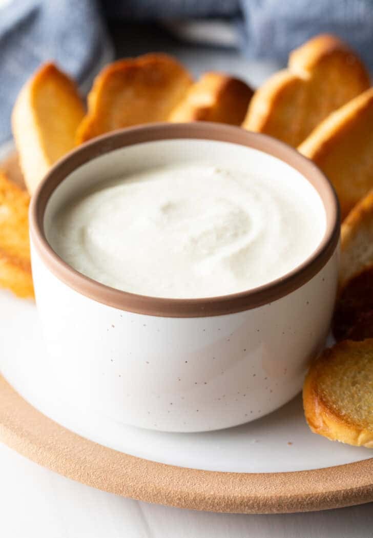 Creamy feta dip in a white bowl, on a plate with slices of toasted bread.