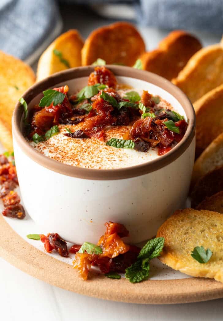 White cup with whipped feta with chopped sun dried tomato, on a plate surrounded by slices of toasted baguette.
