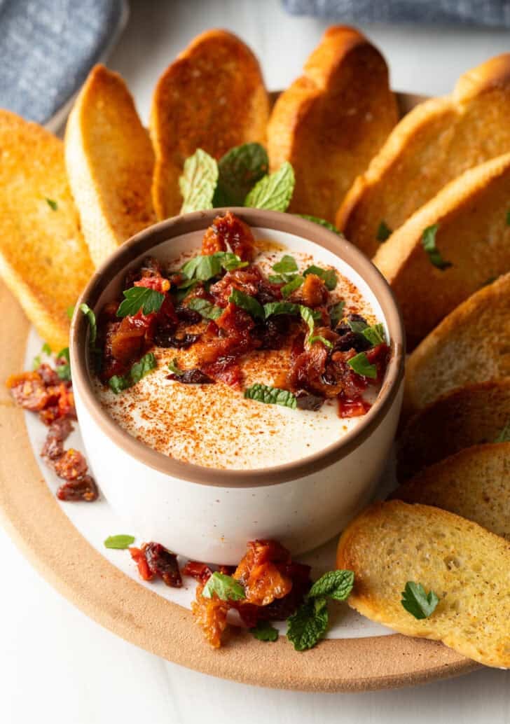 White cup with whipped feta with chopped sun dried tomato, on a plate surrounded by slices of toasted baguette.