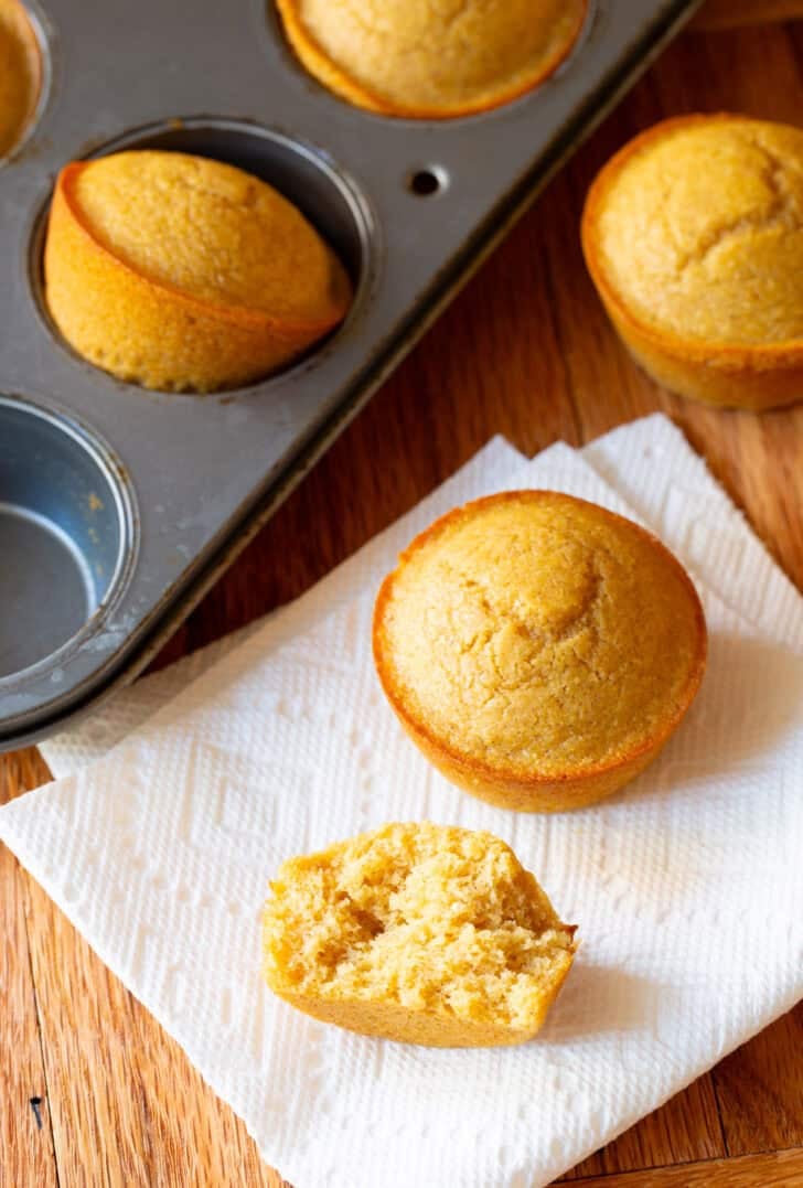 Two honey cornbread muffins on a white napkin, next to a muffin baking tin with more muffins. One muffin has been split in half to show the inside.