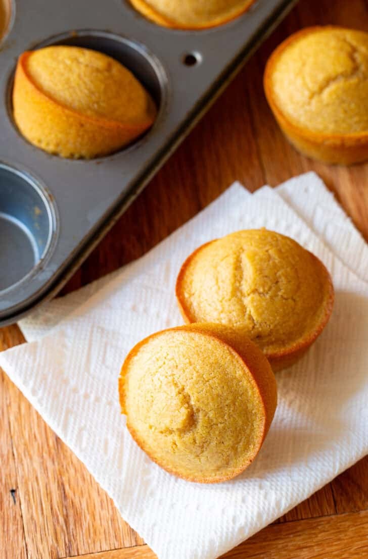 Two honey cornbread muffins on a white napkin, next to a muffin baking tin with more muffins.