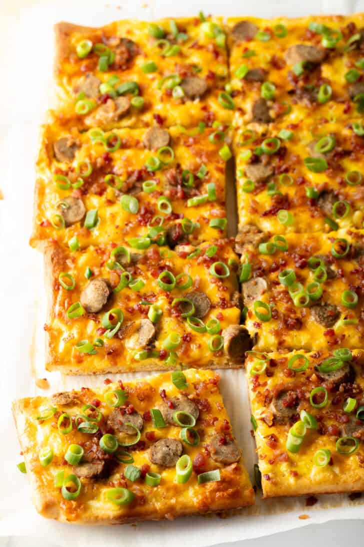 Rectangle baking sheet tray with breakfast pizza, loaded with cheese, sausage, and green onions. The pizza has been cut into 8 rectangles, with one piece pulled away.