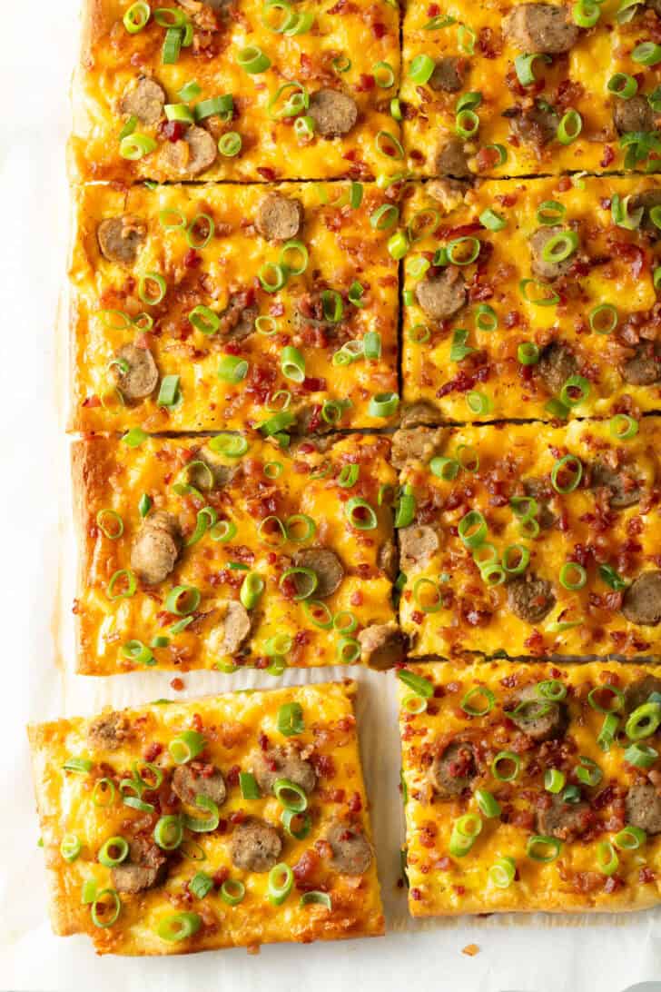 Rectangle baking sheet tray with breakfast pizza, loaded with cheese, sausage, and green onions. The pizza has been cut into 8 rectangles, with one piece pulled away.