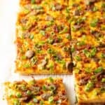 Rectangle baking sheet tray with breakfast pizza, loaded with cheese, sausage, and green onions. The pizza has been cut into 8 rectangles, with one piece pulled away.