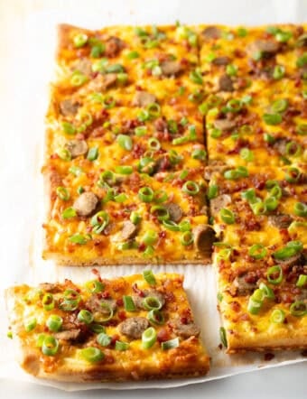 Rectangle baking sheet tray with breakfast pizza, loaded with cheese, sausage, and green onions. The pizza has been cut into 8 rectangles, with one piece pulled away.