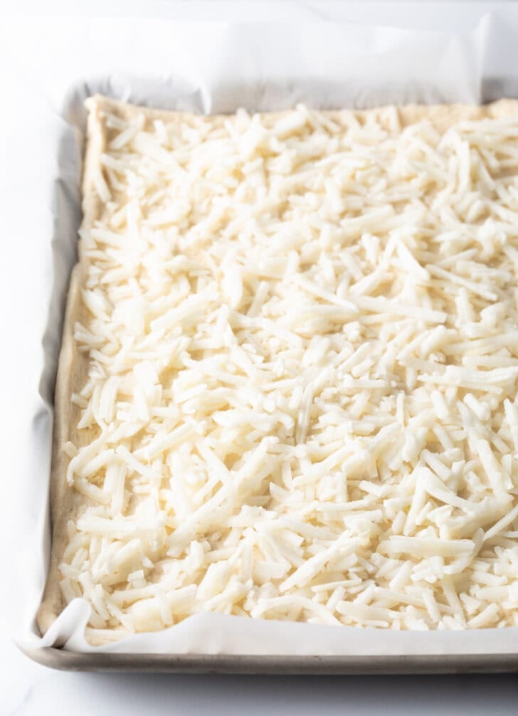 Shredded mozzarella cheese on the top of an unbaked rectangle pizza.