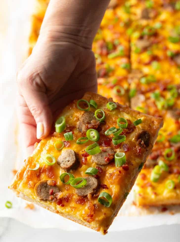 Rectangle baking sheet tray with breakfast pizza, loaded with cheese, sausage, and green onions. The pizza has been cut into 8 rectangles, with a hand pulling one piece away.