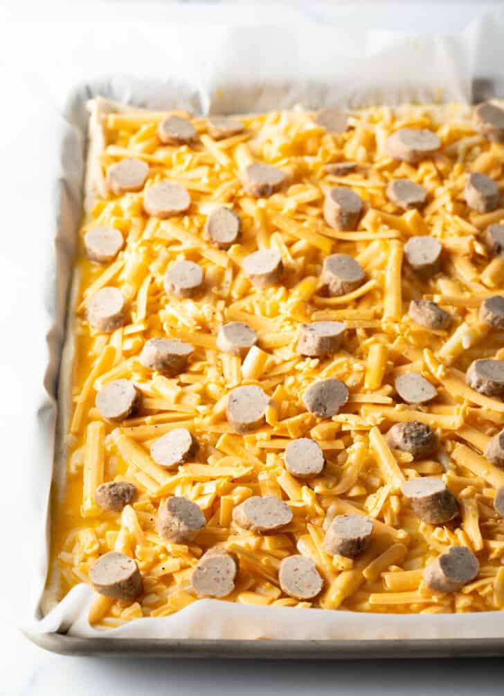 Shredded mozzarella cheese and sliced breakfast sausage pieces on the top of an unbaked rectangle pizza.