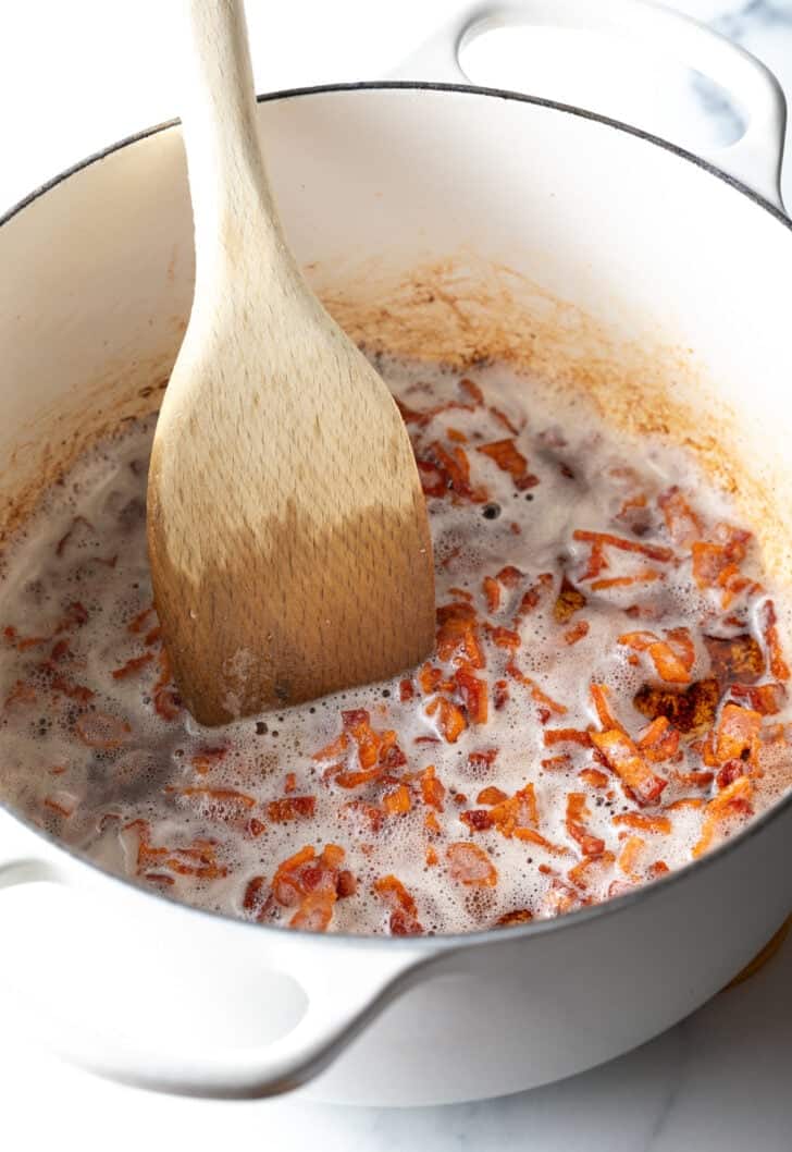 Cooking chopped pieces of bacon in a white cooking pot, being stirred with a wooden spatula.